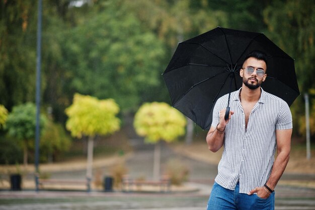 El hombre de barba árabe alto de moda usa jeans de camisa y gafas de sol con paraguas posados bajo la lluvia en la plaza del parque