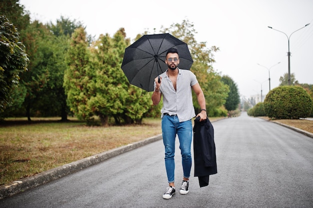 El hombre de barba árabe alto de moda usa pantalones vaqueros y gafas de sol caminando en el parque con paraguas y abrigo a mano