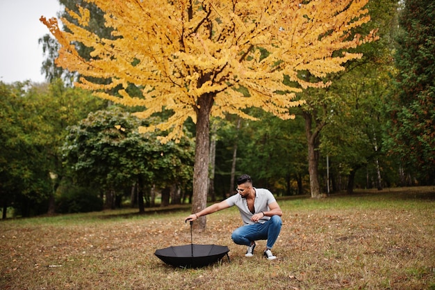 El hombre de barba árabe alto de moda usa pantalones vaqueros y gafas de sol posados en el parque de otoño con paraguas contra el árbol de hojas amarillas