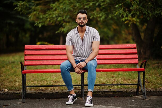 El hombre de barba árabe alto de moda usa pantalones vaqueros y gafas de sol sentados en un banco rojo en el parque