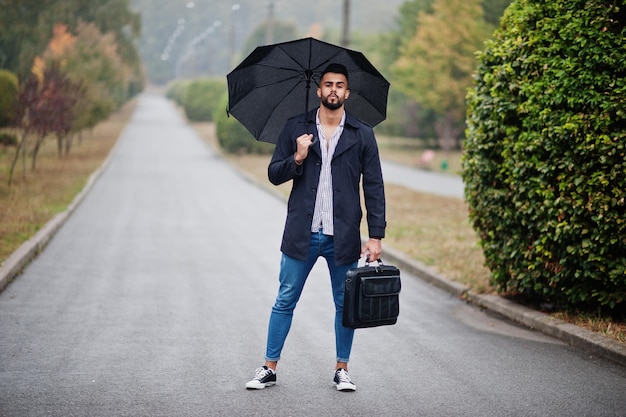 El hombre de barba árabe alto de moda viste un abrigo negro con paraguas y una bolsa que se presenta en el día del clima lluvioso