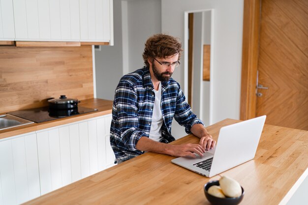 Hombre con barba en camisa a cuadros usando laptop en la cocina.