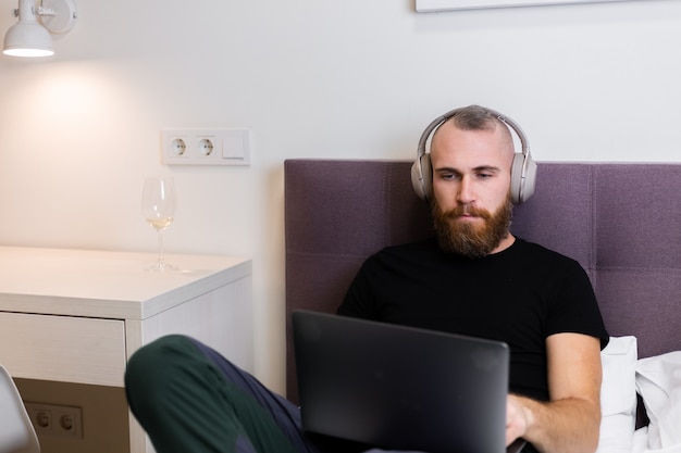 Hombre de barba caucásica en auriculares en el dormitorio en la cama trabajando en la computadora portátil desde casa, escribiendo, pensando.