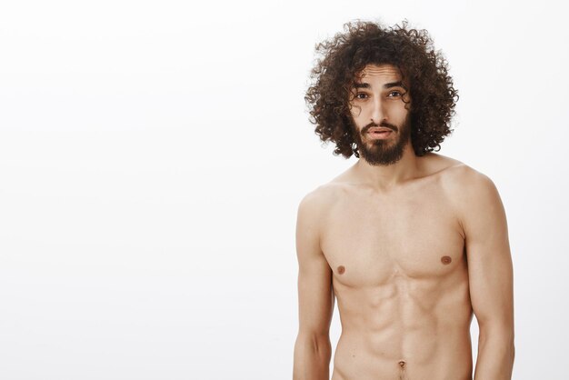 Hombre con barba y corte de pelo rizado que se siente confiado y luciendo sexy con buen cuerpo de pie cerca del espejo en la mañana vistiéndose para trabajar