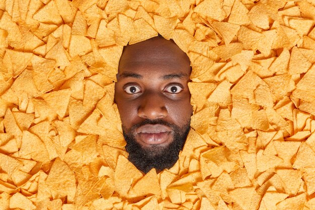 Hombre con barba espesa mira impresionado rodeado de patatas fritas crujientes come bocadillos poco saludables consume muchas calorías
