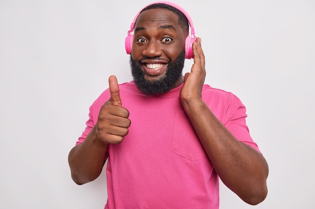 El hombre con barba espesa sonríe ampliamente muestra un signo excelente mantiene el pulgar hacia arriba escucha música en auriculares inalámbricos viste una camiseta rosa sobre blanco