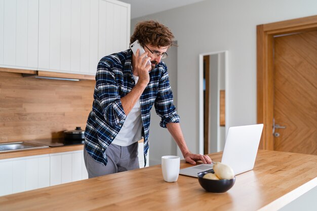 Hombre con barba hablando por teléfono y usando la computadora portátil en la cocina.