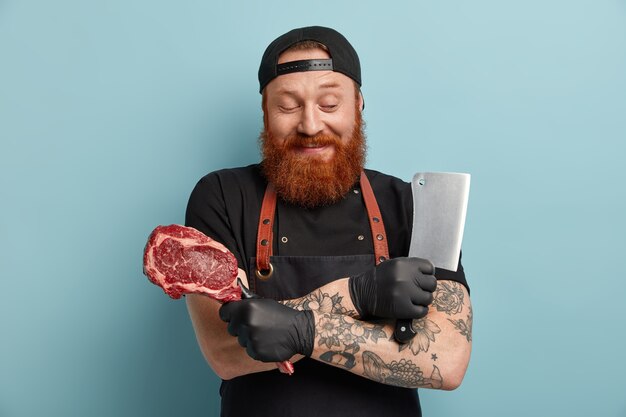 Hombre con barba de jengibre en delantal y guantes con cuchillo y carne