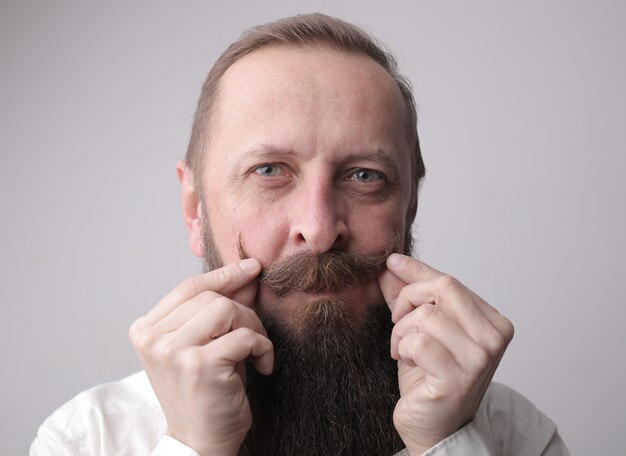 Hombre con barba larga y bigote sonriendo mientras está de pie frente a una pared gris