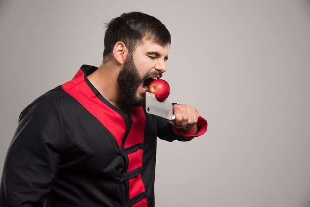 Hombre con barba mordiendo una manzana roja en un cuchillo.