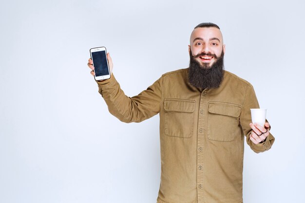 Hombre con barba mostrando su teléfono inteligente mientras sostiene una taza de café.