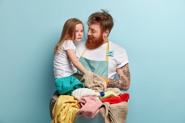 Hombre con barba pelirroja lavando ropa y sosteniendo a su hija