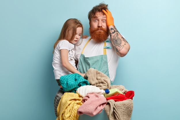 Hombre con barba pelirroja sosteniendo a su hija y lavando ropa