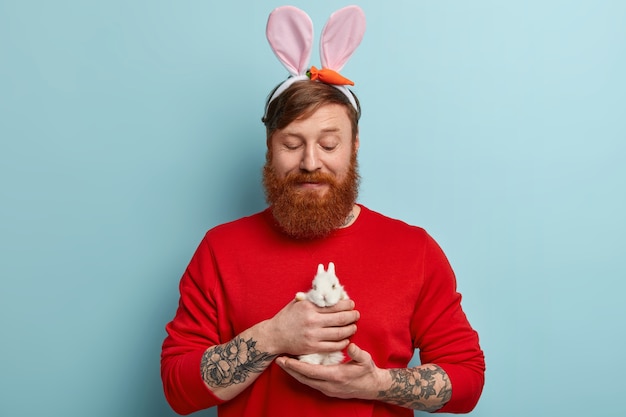 Hombre con barba pelirroja vistiendo ropas coloridas y orejas de conejo sosteniendo conejito