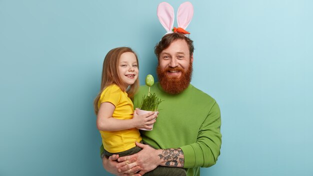 Hombre con barba pelirroja vistiendo ropas coloridas y orejas de conejo sosteniendo a su hija