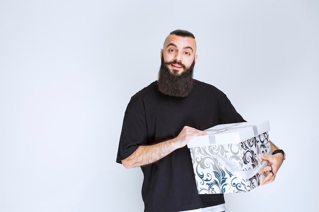 Hombre con barba sosteniendo una caja de regalo azul blanca y parece sorprendido.