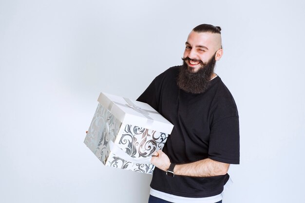 Hombre con barba sosteniendo una caja de regalo azul blanca sonriendo y sintiéndose feliz.