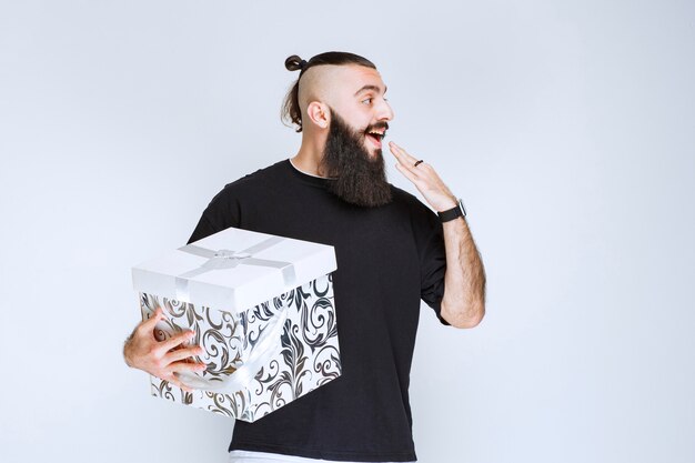 Hombre con barba sosteniendo una caja de regalo azul blanca sonriendo y sintiéndose feliz.