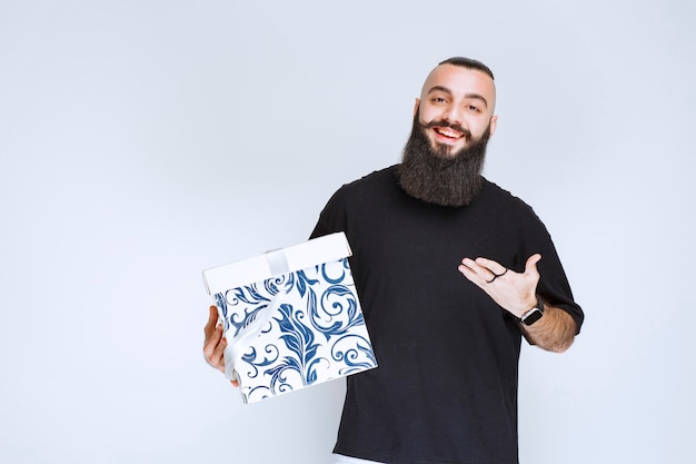 Hombre con barba sosteniendo una caja de regalo azul blanca sonriendo y sintiéndose feliz.