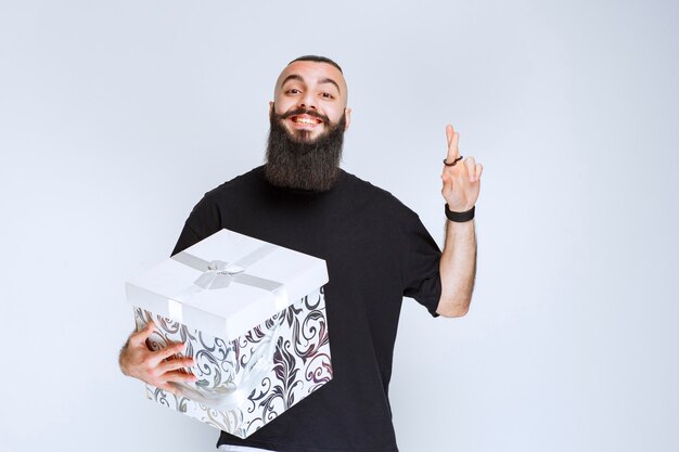 Hombre con barba sosteniendo una caja de regalo azul blanca sonriendo y sintiéndose feliz.