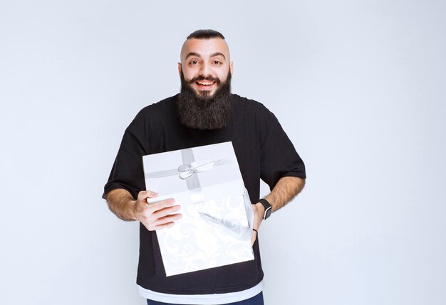 Hombre con barba sosteniendo una caja de regalo azul blanca sonriendo y sintiéndose feliz.