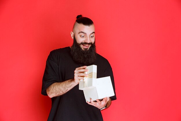 Hombre con barba sosteniendo una caja de regalo blanca y abriéndola con entusiasmo.