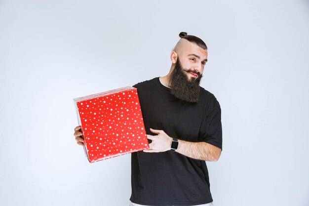 Hombre con barba sosteniendo una caja de regalo roja y parece cansado.