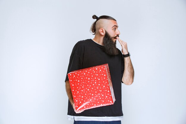 Hombre con barba sosteniendo una caja de regalo roja y parece confundido y aterrorizado.