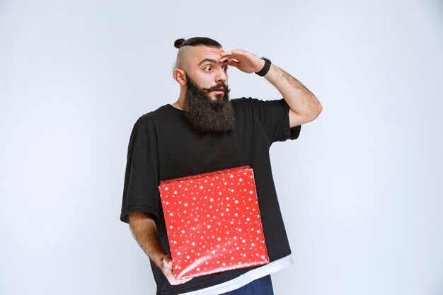 Hombre con barba sosteniendo una caja de regalo roja y parece confundido y aterrorizado.