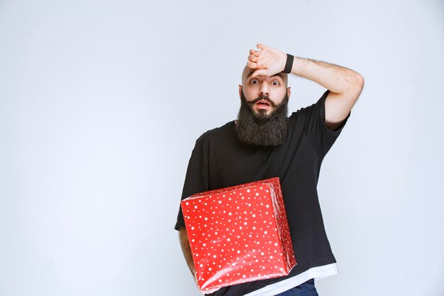 Hombre con barba sosteniendo una caja de regalo roja y parece confundido y aterrorizado.