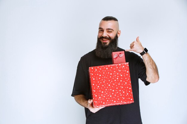 Hombre con barba sosteniendo cajas de regalo rojas y apuntando hacia atrás.