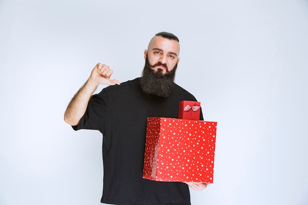 Hombre con barba sosteniendo cajas de regalo rojas y apuntando la parte trasera con una cara pálida.