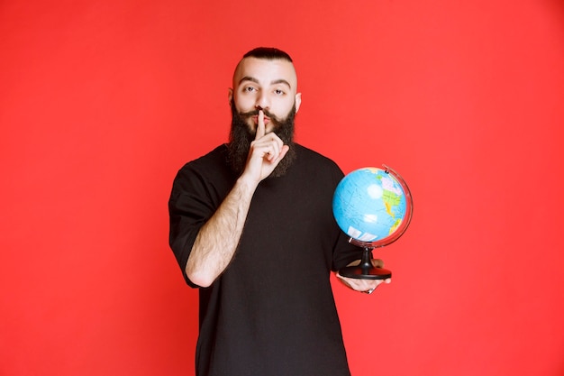 Hombre con barba sosteniendo un globo terráqueo y pidiendo silencio.