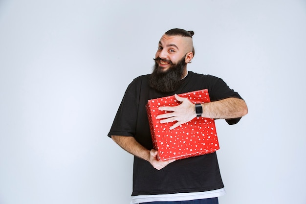 Hombre con barba sosteniendo su caja de regalo roja, disfrutándolo y sintiéndose feliz.