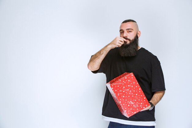 Hombre con barba sosteniendo su caja de regalo roja, disfrutándolo y sintiéndose feliz.