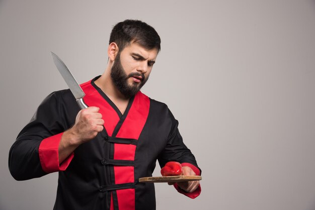 Hombre con barba tratando de cortar un pimiento rojo sobre tabla de madera.