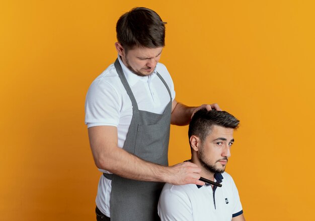 Hombre barbero en delantal afeitarse la barba con navaja de cliente satisfecho sobre pared naranja