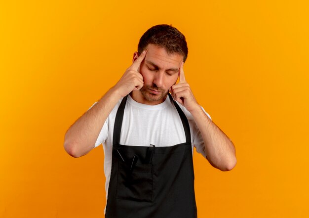 Hombre barbero en delantal apuntando sus sienes con aspecto cansado y aburrido de pie sobre la pared naranja