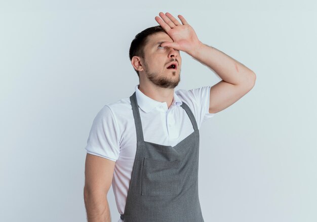 Hombre barbero en delantal con aspecto cansado y aburrido con las manos sobre la cabeza rodando los ojos de pie sobre la pared blanca