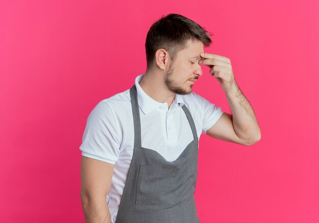 Hombre barbero en delantal con aspecto cansado y con exceso de trabajo tocando la nariz entre los ojos cerrados de pie sobre la pared rosa