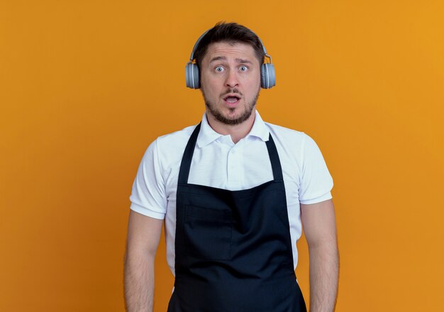 Hombre barbero en delantal con auriculares mirando canmera confundido y muy ansioso de pie sobre la pared naranja