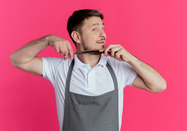 Hombre barbero en delantal cortando su barba con unas tijeras de pie sobre una pared rosa