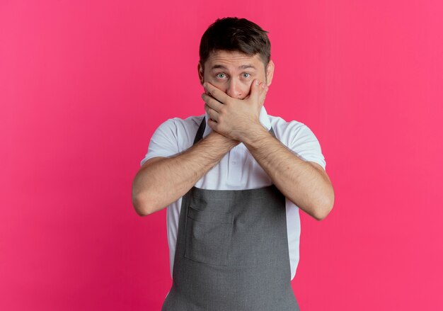 Hombre barbero en delantal cubriendo la boca con las manos en estado de shock de pie sobre la pared rosa