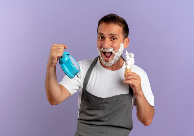 Hombre barbero en delantal con espuma de afeitar en su rostro sosteniendo reloj despertador y brocha de afeitar mirando sorprendido de pie sobre la pared púrpura