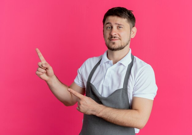 Hombre barbero en delantal con expresión de confianza apuntando con el dedo índice hacia el lado parado sobre la pared rosa