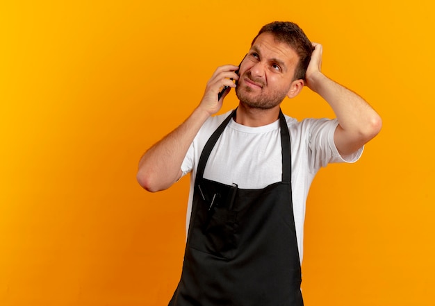 Hombre barbero en delantal hablando por teléfono móvil mirando disgustado parado sobre pared naranja
