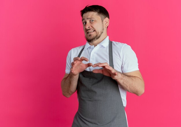 Foto gratuita hombre barbero en delantal haciendo gesto de defensa sosteniendo las manos con expresión de disgusto de pie sobre la pared rosa