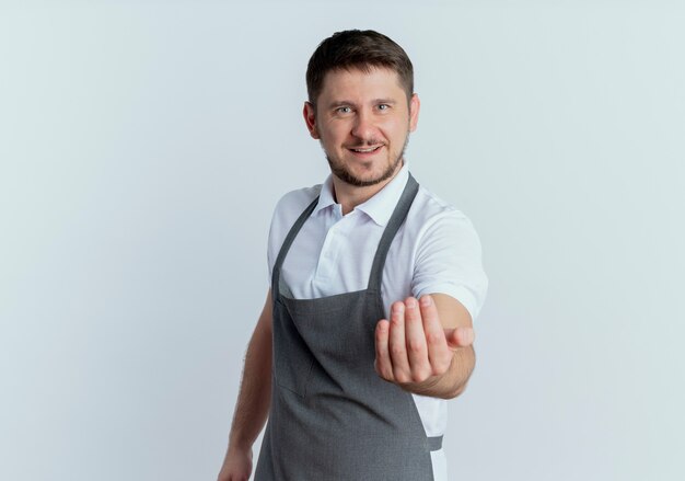 Hombre barbero en delantal haciendo gesto de ven aquí con la mano sonriente de pie amistoso sobre fondo blanco.