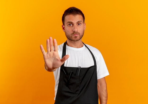 Hombre barbero en delantal haciendo señal de stop con la mano mirando hacia el frente con cara seria de pie sobre la pared naranja
