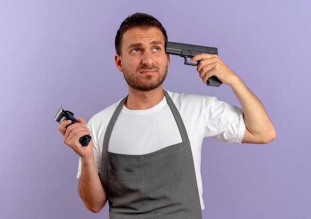 Hombre barbero en delantal manteniendo la pistola cerca de su sien mirando disgustado parado sobre la pared azul